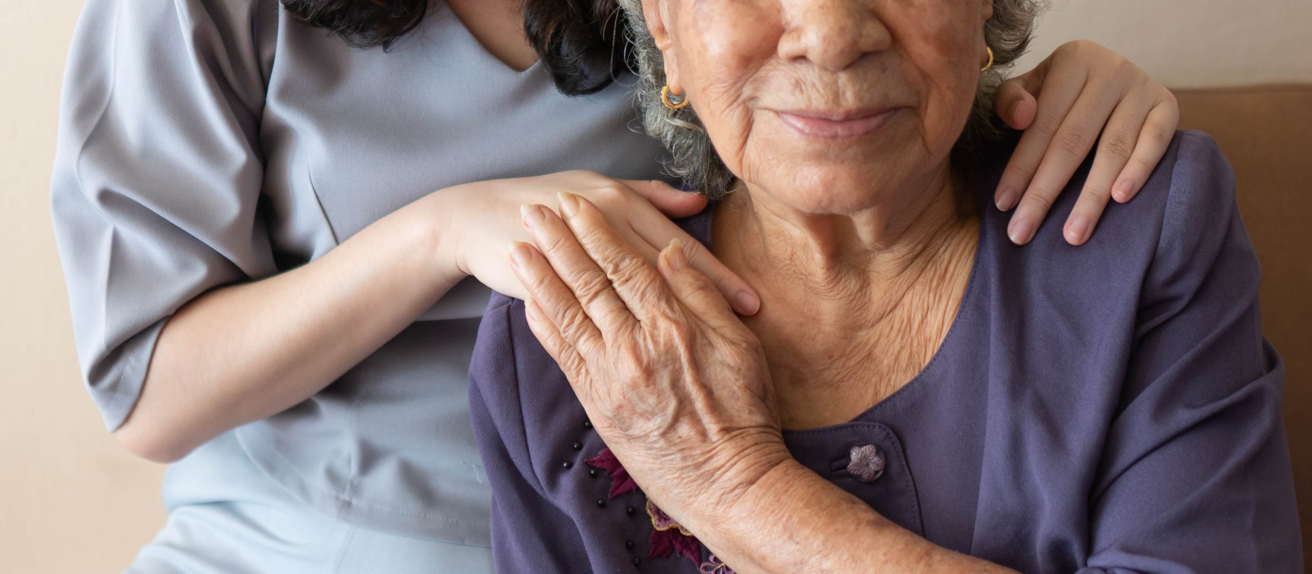 A young woman tenderly puts her hands on the shoulders of an older woman, who holds one of her hands. Faces not pictured entirely.